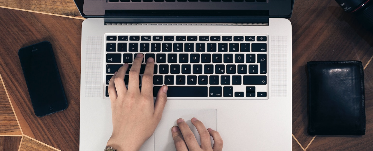 Mão masculina pesquisando em um notebook como investir e quais tipos de investimentos combinam com seus objetivos, acompanhado de seu celular ao lado direito e carteira ao lado esquerdo